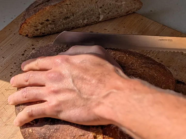 Mà que subjecta un pa de crosta fosca mentre es talla amb un ganivet de pa